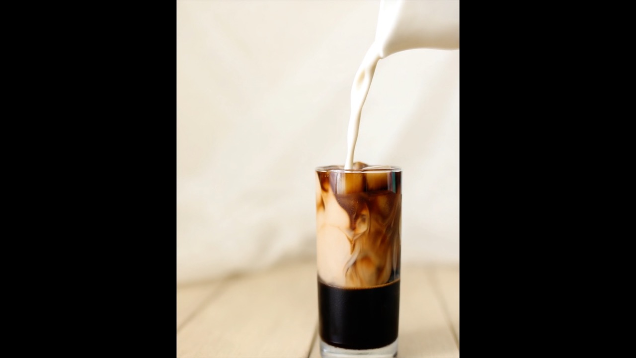 Milk pouring into iced coffee, slow motion, creamy swirl, warm natural light